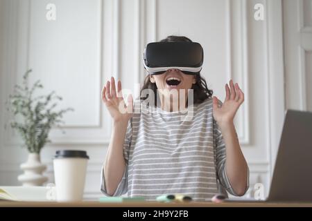 Excited entrepreneur lady tests virtual reality technology wearing VR glasses sitting at home office Stock Photo