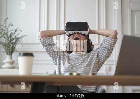 Amazed woman office employee in VR glasses laughing and leaning back in chair with hands behind head Stock Photo