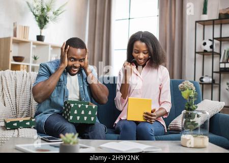 African american man and woman with real happiness on their face opening colorful gift boxes. Pleasant young couple sharing with presents while sitting on comfy couch. Stock Photo