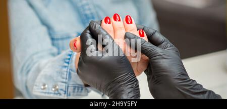 Examination of manicured fingernails. Hands of manicure master in black gloves examining female red nails in manicure salon Stock Photo