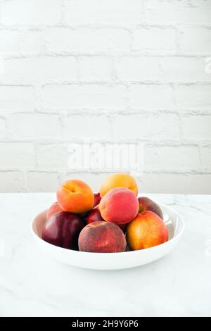 Whole peaches, nectarines and apricots in a white porcelain bowl on white marble table with brick wall in the background. Low angle view and copy spac Stock Photo