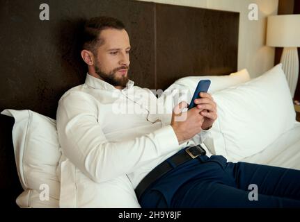 Handsome young European bearded man, successful businessman browsing and chatting on mobile phone, lying on a bed in the bedchamber of a hotel, relaxi Stock Photo