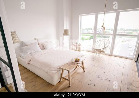 Large Bright Bedroom in a Minimalist Style in Pastel Cream Colors with a Double Bed, White Walls, a Wicker Laundry Basket, Wicker Chandeliers, Beige Bed Bench, Large Window , Boho Hanging Chair and Wooden Floor. . High quality photo Stock Photo