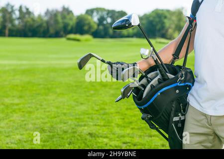 hand in a black leather glove pulls a golf club out of a bag. Bag full of golf clubs, iron, wedge, wood, drivers. Course of Turf Grass Background. Copy Space. Close-up. . High quality photo Stock Photo