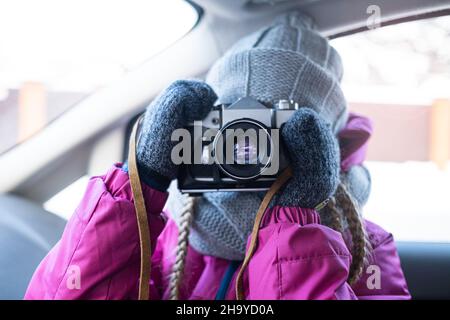 A girl in winter clothes takes a picture with a camera while in the car. Road travel in winter by car. Vintage film SLR camera on a leather strap in Stock Photo