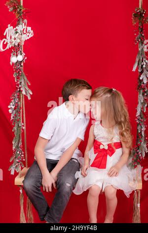 boy and girl - sister and brother - sitting on a swing decorated with christmas decor and tree branches. on red background. siblings on new year. Stock Photo