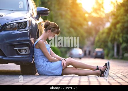 Young woman driver sitting beside her broken car waiting for help. Vehicle problems concept. Stock Photo