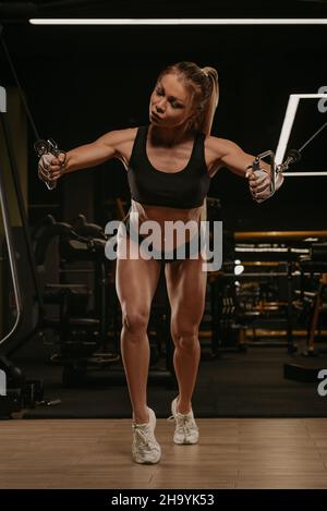 A close-up photo of a fit woman with blonde hair who is doing a chest  workout on the cable machine in a gym. A girl is training her pectoral  muscles Stock Photo 
