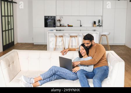 Cheerful couple in love spending leisure time online with a laptop at home. Young woman and man looking at the laptop screen while sitting on the sofa, watching comedy movies, funny videos Stock Photo