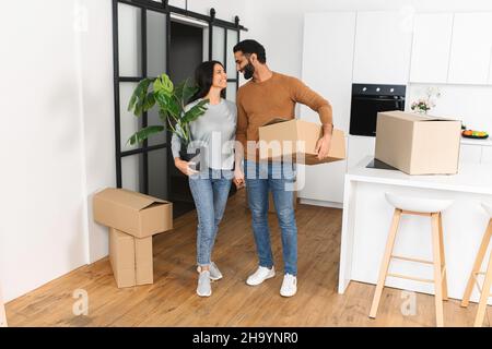 Attractive couple wearing casual clothing moving into new home. Happy man carrying cardboard box and bonding to woman who holding plant. Real estate rent, mortgage and ownership concept Stock Photo
