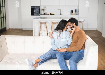 Gentle and lovely couple in love sitting in embrace on the comfortable sofa at home, looking to each other tenderly. Concept of support and caring in marriage Stock Photo