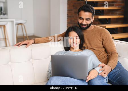 Young Pakistani couple using laptop while sitting on the sofa at home. Spouses watching movie on laptop. Happy man and woman embracing while relaxing on couch Stock Photo