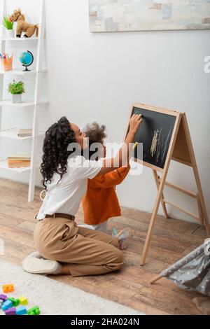 Side view of african american woman and kid drawing on chalkboard in living room Stock Photo