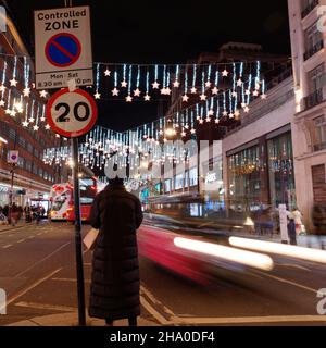 London, Greater London, England, December 04 2021: Lady stands in Oxford Street as the traffic whizzes by at night with Christmas lights on display. Stock Photo