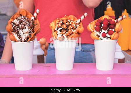 Bubble waffles with raspberries, chocolate and marshmallow in white paper cups are sold on food festival event. Street sweet food in food stall. Stock Photo