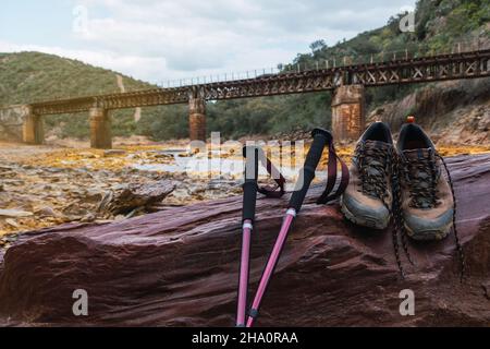Hiking boots and trekking poles on a focused stone and in the background an old iron bridge over the Rio Tinto. Stock Photo