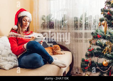 Young woman writing goals list for 2022 New year in notebook at home sitting by Christmas tree wearing Santa hat and warm sweater. Stock Photo