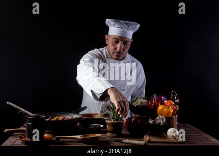 Male cook in white uniform and hat while working in a restaurant kitchen. On the table vegetables and herbs ingredients Stock Photo