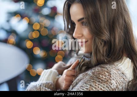 Portrait of happy young attractive brunette woman in cozy knitted sweater resting at home near Christmas tree. Concept of enjoying free time with good mood in Christmas atmosphere. Stock Photo