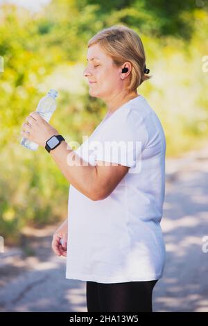 Side view photo of a blonde woman outdoors wearing sports clothes having digital watch on her hand and headphones drinking some water from the plastic Stock Photo
