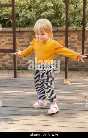 A beautiful cheerful little girl in a stylish striped pantsuit dancing in a city park. Lifestyle Stock Photo