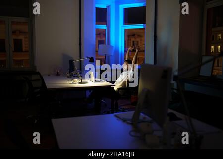 Remote programmer work late night stretching hands while sitting at laptop computer in dark office Stock Photo