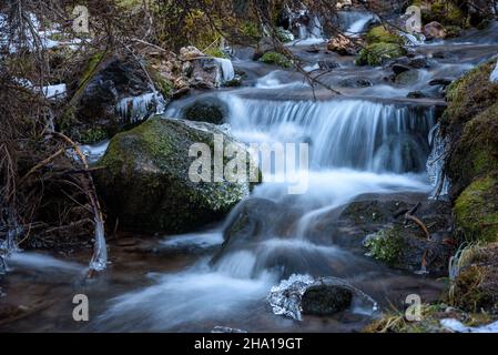 Small waterfall in a snowy forest in the mountains on a cold winter day. Icy rocks are in foreground. Stock Photo
