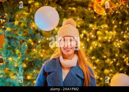 A smiling girl stands against the background of large glowing Christmas tree. Selective focus. Festive mood Stock Photo
