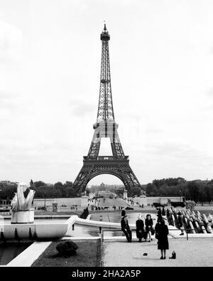 Paris Eiffel Tower 1945 from Trocadero Gardens. Full height view of the tower taken from above the fountain of the Jardins du Trocadero (Gardens of the Trocadero) The camera position is slightly above and perhaps 20 feet away from a family of eight. A woman in a dress appears to be arranging a photo of some of the family with the tower in the background. The family group in the foreground appear to age from 5 to 80 years. Below them, there are many people of foot at the base of the fountain and walking over Pont d’lena. Stock Photo
