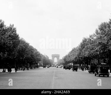 Long view up the Champs-Élysées with the Arc de Triomphe in the distance. The camera position is standing in the middle of the Avenue des Champs-Élysées along the center line. The location is perhaps near the present-day Square Berlin looking northwest nearly 1.5 km to the Arc de Triomphe. The few automobiles near the camera position include military jeeps. Heavier automobile traffic is apparent in the distance. There are bicyclists and pedestrians on either side of the boulevard. The lower third of the image is primarily empty grey pavement. The upper third of the image is a “V” of white sky Stock Photo