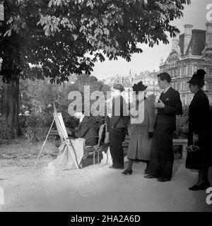 Paris scene painter with observers, 1945, near Place du Carrousel. A painter is seated in front of a canvas on a easel stand.He is dressed in suit and hat and he holds a cigarette from his mouth. It is not apparent what he is painting. He is facing away from what appears to be buildings of the Louvre and sits in park space at the base of a mature tree. The background to the left of the image is a tangle of park-like greenery. Four intersting figures are standing as if in a queue and staring intently over the painter’s left shoulder at his canvas. Stock Photo