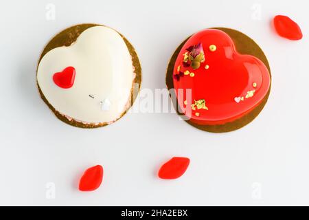 A cake in heart shape. Two heart shaped mousse cupcakes white and red  and candy on a white background. Valentine's Day. Stock Photo