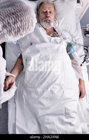 Top View On Elderly Sick Ill Patient Male Lying On Bed With Endotracheal Tube, Female Doctor Checking The Pulse. Coronavirus, Covid-19.Treatment, Medi Stock Photo