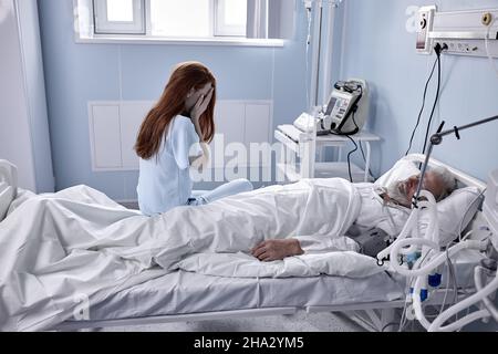 In Hospital, Worrying Young Mistress Sits Beside the Bed where Sick Man Lies. Redhead Woman Is Crying, Hopes for Recovery. In medical Ward Room. Healt Stock Photo