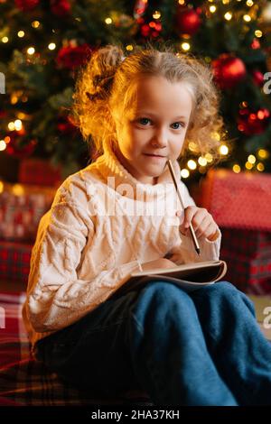 Vertical shot of pretty thinking little blonde girl writing letter to Santa Claus sitting on floor on background of Christmas tree and gift boxes. Stock Photo
