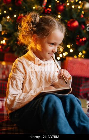 Vertical shot of charming dreaming little blonde girl writing letter to Santa Claus sitting on floor on background of Christmas tree and gift boxes. Stock Photo