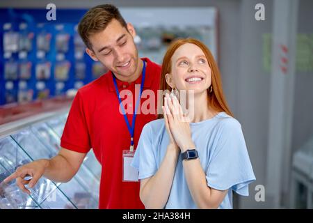 Positive Redhead Lady In Casual Outfit Stand By Shelves With Smartphones In Store, With Consultant, Cute Customer Woman Is Dreaming About New Modern P Stock Photo