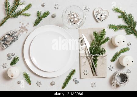 Christmas table setting with silver decorations and fir branches on a gray background. Empty white plate for design. Top view, flat lay. Stock Photo