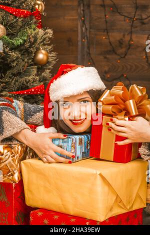 Lovely Young Woman in a Red Santa Hat Holding a Dozen of Christmas Multi Colored Gifts. Decorated Christmas Tree, Electric Garland on the Wall, Lights. Close-up. Wood Background. High quality photo Stock Photo