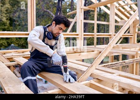 Hardworking Caucasian Contractor Working With Drill Driver. Focused Carpenter Working on Wooden House Skeleton Frame Roof Section. Construction Indust Stock Photo