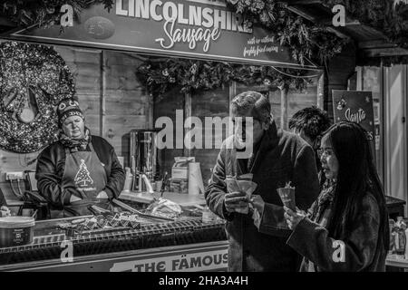 Harrogate couple eating Lincolnshire sausages and other fresh food at a market stall as the vendor watches on, North Yorkshire, England, UK. Stock Photo