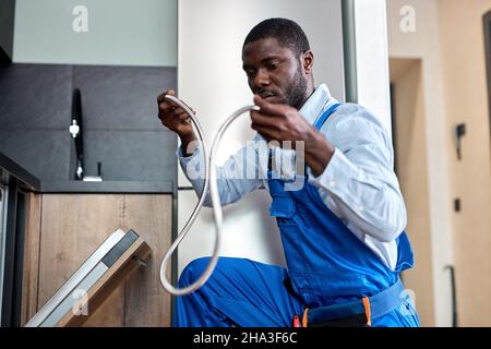 Young African Handyman Repairing Dishwasher, Need To Change Old Dishwasher Hose, Black Guy In Blue Overalls Workwear Is Concentrated On Work, In Kitch Stock Photo