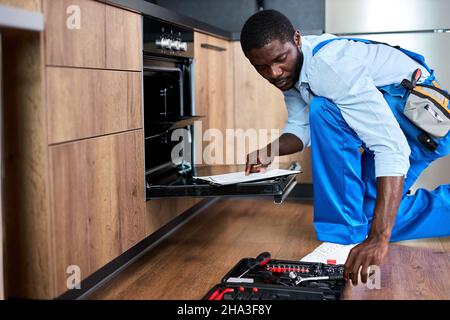 Don't delay with repair. Side view afro repairman examining oven in kitchen with tool case. Confident professional black repairman handyman in blue wo Stock Photo