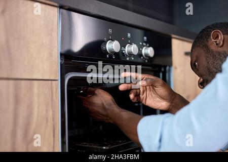 Hardworking afro american guy in blue workwear repairing broken electric oven in kitchen at home indoors, using worktool. side view portrait. handyman Stock Photo