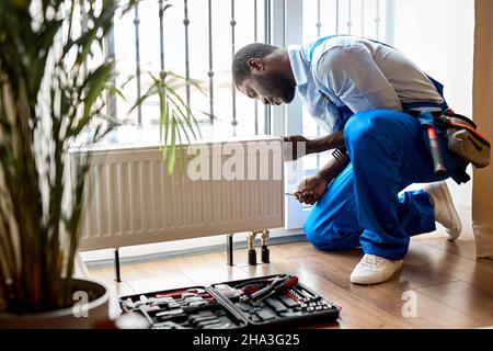 afro repairman in blue overalls using tools while installing or repairing heating radiator. plumber installing heating system in apartment. radiator i Stock Photo