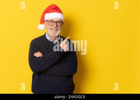 Senior smiling man wearing santa hat pointing finger up standing isolated over yellow background. Stock Photo