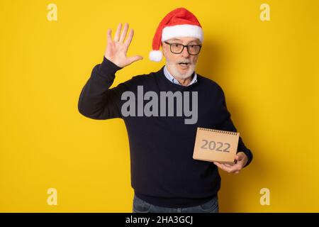 Senior excited man wearing santa hat showing calendar isolated over yellow background. Stock Photo