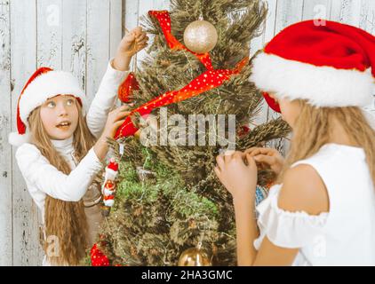 Christmas girls decorate the Christmas tree together with decorative elements. Two sisters hang decorations on a Fir tree in their living room at home. Wooden Background. Close-up. High quality photo Stock Photo