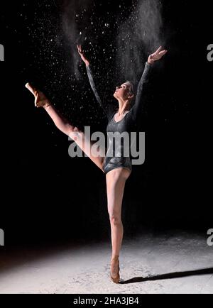 graceful slim woman in black bodysuit emotionally dancing on black background in clouds of white dust, in studio. ballerina female in slow motion acti Stock Photo
