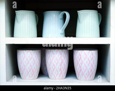 Three light blue pitchers are displayed on the top shelf of a white shelf unit. Three pink and white patterned mugs are on the bottom shelf. Stock Photo
